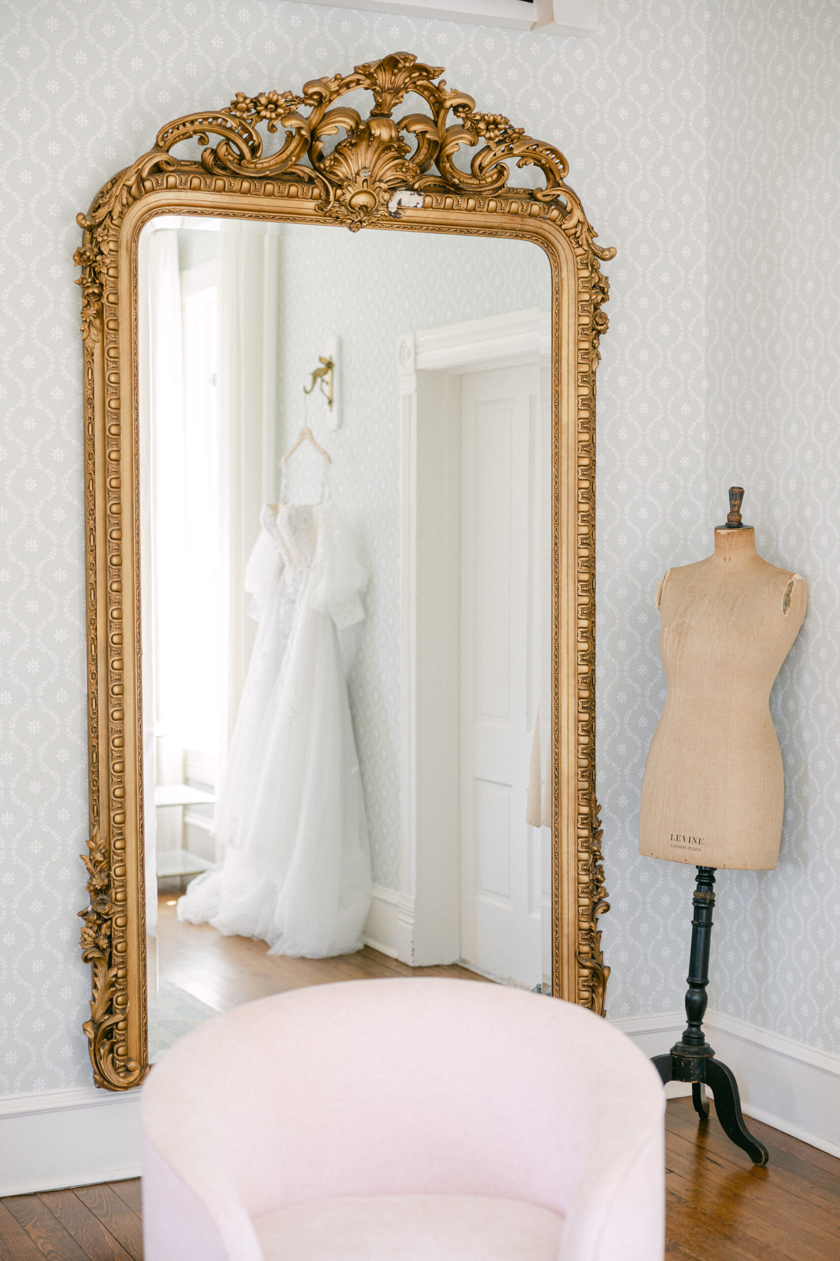 Elegant bridal suite at Woodbine Mansion featuring a gold ornate mirror reflecting a hanging lace wedding gown, a vintage dress form, and pastel decor.